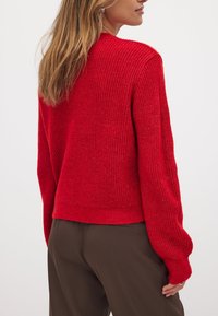 Roter gerippter Pullover mit lockerer Passform und langen Ärmeln, der ein strukturiertes Strickmuster aufweist und in einer gekürzten Länge auf der Taille endet.