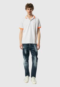 Polo bianco con colletto e dettagli arancioni sulle maniche, abbinato a jeans in denim scuro con particolari scoloriti. Il modello si trova in un ambiente neutro.