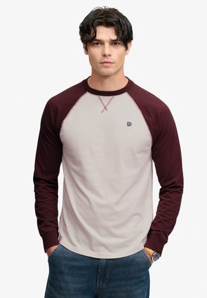 Junger Mann mit dunklen Haaren trägt ein langärmeliges Raglan-Shirt mit burgunderfarbenen Ärmeln und einem hellbeigen Körper, steht mit den Händen in den Taschen.