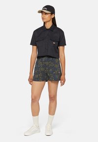 Gekürztes schwarzes Hemd mit zwei Brusttaschen, kombiniert mit gemusterten Shorts in Blau und Grün. Weiße Sneaker und hellfarbige Socken.