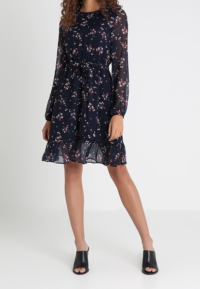 Femme portant une robe fleurie bleu marine avec des manches longues en voile transparent, une ceinture cintrée à la taille, un ourlet à volants et des mules à talons ouvertes noires.