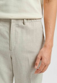 Ljust beige linnebyxor med en slät textur, som har en sidficka och en midjeband för passformjustering. Designen är skräddarsydd och minimalistisk.