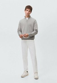 Sudadera gris con cremallera, de tela suave, modelo de pie con pantalones blancos y zapatillas claras. Diseño minimalista con una paleta de colores neutros.