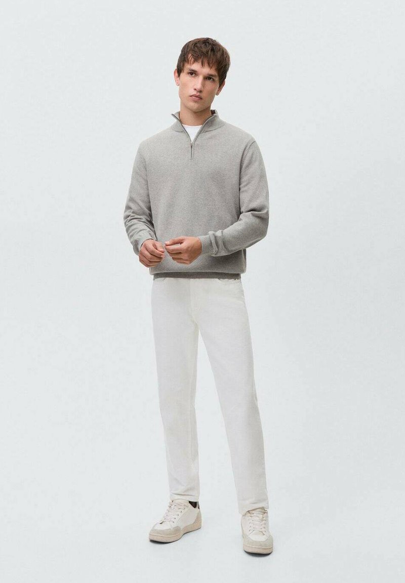 Sudadera gris con cremallera, de tela suave, modelo de pie con pantalones blancos y zapatillas claras. Diseño minimalista con una paleta de colores neutros.