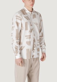 Camisa de algodón ligera con una base blanca que presenta patrones abstractos en beige. Mangas largas, cierre de botones en la parte delantera y cuello clásico.