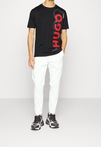 Mand iført sort T-shirt med stor rød "HUGO" tekst, hvide bukser og sort-hvide sneakers, stående med den ene hånd i lommen.