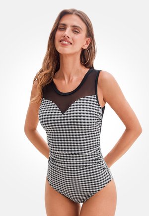 Kobieta ubrana w czarno-białe body w kratę bez rękawów z przezroczystą czarną siateczką przy dekolcie, stojąca z rękami za plecami.