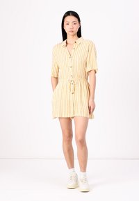 Robe courte rayée jaune avec un devant à boutons, des manches courtes et une taille à cordon. Portée avec des baskets blanches et des chaussettes mi-mollet.