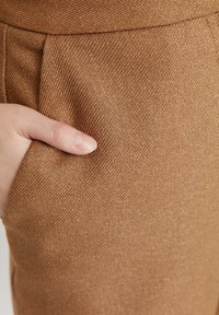 Hnědá texturovaná tkanina s jemným diagonálním vzorem, která obsahuje boční kapsu a umístění ruky v kapse. Materiál se zdá být měkký.