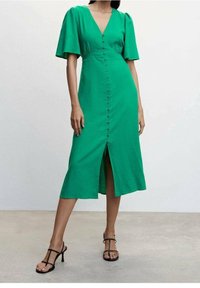 Robe verte jusqu'aux genoux avec col en V, manches courtes bouffantes et une fente à l'avant. Dotée d'une rangée de boutons au centre. Tissu lisse.