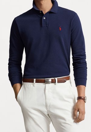 Uomo che indossa una polo a maniche lunghe blu navy infilata in pantaloni bianchi con cintura in pelle marrone e cinturino dell'orologio abbinato.