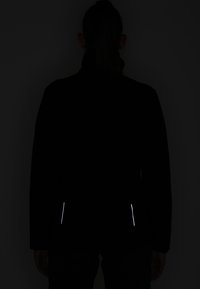 Veste en polaire noire avec un col montant, dotée de zips réfléchissants à la taille. Le matériau semble doux avec une texture lisse.