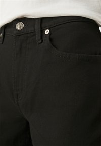 Jeans de denim negro con un diseño clásico de cinco bolsillos, que presentan un botón plateado y costuras en contraste. Textura suave, ajuste entallado.