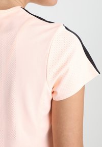 Sporttopp med kort ärm i ljusrosa tyg med svarta detaljer, med ett strukturerat mönster och perforeringar för andningsförmåga.
