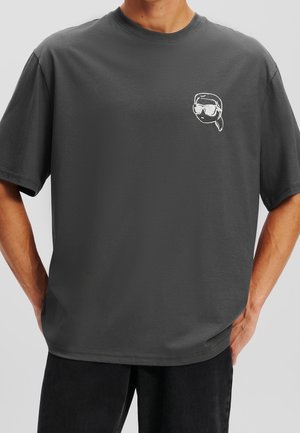 Homme portant un t-shirt gris foncé avec un petit graphique blanc représentant un visage portant des lunettes de soleil et un bandana sur la poitrine gauche.