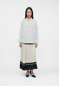 Camisa de botones gris claro con botones dorados, combinada con una falda crema con pliegues que tiene un borde negro y detalle en texto, complementada con tacones negros.