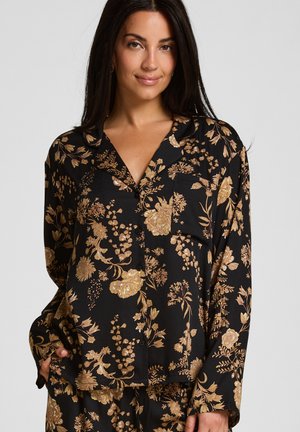 Femme portant un chemisier noir boutonné avec des motifs floraux dorés et des manches longues, posant avec une main dans la poche.