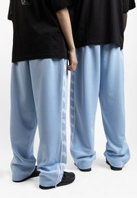 Light blue, loose-fitting track pants with side zippers and white logo accents. Smooth texture, paired with black oversized t-shirts.
