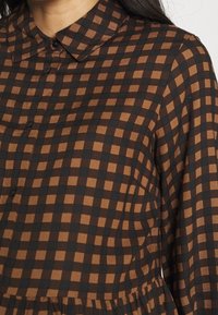 Robe en tissu à carreaux marron et noir avec un col, présentant une fermeture boutonnée à l'avant et une taille froncée, vue de la poitrine vers le haut.