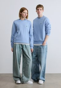 Ανοιχτό μπλε φούτερ με πράσινα λογότυπα, συνδυασμένα με φαρδιά ανοιχτόχρωμα τζιν. Άσπρα αθλητικά παπούτσια ολοκληρώνουν το casual look.