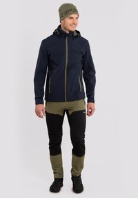 Marineblauwe softshell jas met een volledige rits, groene accenten en twee zijzakken. Gecombineerd met aansluitende olijfgroene en zwarte broeken en zwarte wandelschoenen.