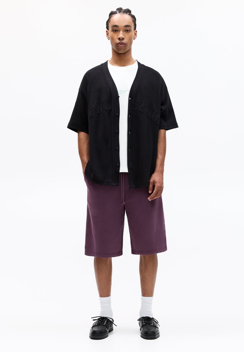 Schwarzer oversized Cardigan mit vorderen Knöpfen und geprägtem Text, weißes T-Shirt, lila Shorts, weiße Socken und schwarze Schuhe.
