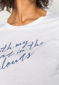 Camiseta de algodón blanca con cuello redondo, que presenta letras en cursiva de color azul marino "Con la cabeza en las nubes" en la parte frontal.