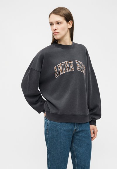 Moteris su tiesiais rudais plaukais, vilkinti tamsiai pilką platų džemperį su užrašu „ANINE BIRG“ ir mėlynais džinsais, stovinti prie balto fono.