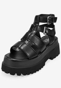 Sandalia negra con plataforma gruesa, con tiras trenzadas y hebillas ajustables. Fabricada en material sintético suave con una suela texturizada.