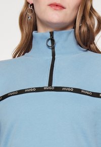 Vrouw in een lichtblauwe rits-top met een zwarte HUGO logo band over de borst en zilveren ringoorbellen met hangende balletjes.