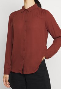 Blouse bordeaux à manches longues avec une fermeture boutonnée à l'avant, un col, et des détails discrets de plis couchés sur les épaules. Fabriquée en tissu lisse.