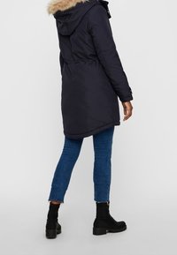 Person med en svart parka med pälsfoder på huvan, blå cropped jeans och svarta klackstövlar, som står mot en enkel bakgrund.