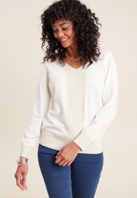 Maglione chiaro in crema con scollo a V testurizzato, maniche lunghe e orlo a coste; presenta un pannello centrale aderente di color crema. Indossato con jeans blu.