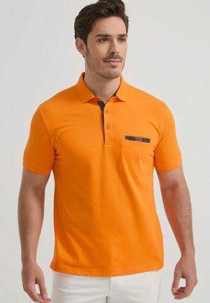 Homme portant un polo orange vif à manches courtes avec des détails foncés et un pantalon blanc, debout devant un fond clair uni.