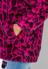 Chaqueta de piel sintética con estampado de leopardo rosa y textura suave, que cuenta con un bolsillo frontal y grandes manchas negras, combinada con unos vaqueros deshilachados.