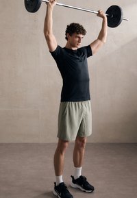 Maglietta sportiva nera, pantaloni corti verde chiaro, mentre solleva un bilanciere. Presenta maniche corte, tessuto leggero e cuciture a contrasto.