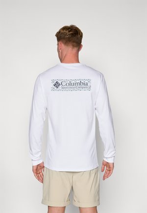 White long-sleeve shirt featuring a large blue graphical logo with patterns on the back. Worn with light beige shorts, simple design.