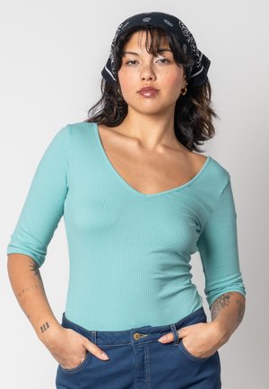 Helles türkisfarbenes, geripptes Langarmoberteil mit einem tiefen V-Ausschnitt, kombiniert mit dunkelblauen Jeans. Das Model trägt ein schwarzes Halstuch und hat sichtbare Tattoos.