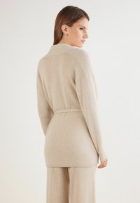 Cárdigan de punto beige con cintura ceñida, mangas largas y una textura suave. Presenta un cuello clásico y acentos de ribete en el dobladillo.