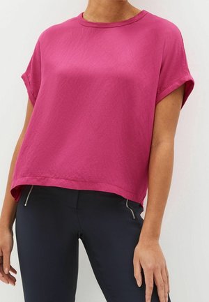 Femme portant un haut ample à manches courtes magenta associé à un pantalon noir ajusté avec des poches zippées à l'avant.