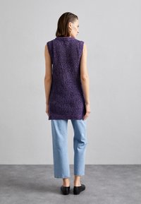 Adīts garš purpura veste ar teksturētu rakstu, bez piedurknēm, un dalītu apakšmalu, kombinēts ar gaiši ziliem platām kājām džinsiem un melniem apaviem.