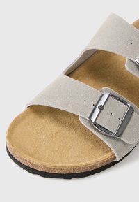 Hellgraue Wildledersandale mit zwei breiten Riemen, silberner Schnalle, Kork-Fußbett und schwarzer Außensohle, von vorne schräg oben gezeigt.