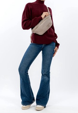 Osoba ubrana w bordowy sweter w warkocze, szerokie niebieskie dżinsy, beżowe trampki oraz beżową torbę na ramię z widocznym zamkiem błyskawicznym i logo.