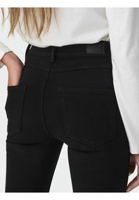 Ženska nosi črne hlače z visokim pasom z zadnjimi žepi, ena roka je v žepu, na sebi ima belo dolgolasno majico, ki je vsulana.