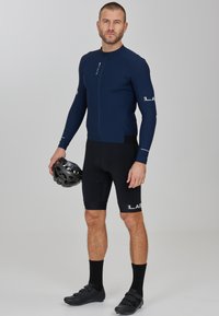 Marineblaues Fahrradtrikot mit langen Ärmeln, schwarze gepolsterte Shorts, schwarze Fahrradschuhe und einen mattschwarzen Helm, der in einer Hand gehalten wird.