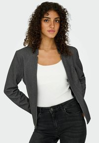 Jeune femme aux cheveux bouclés portant un blazer gris sur un haut blanc et un jean noir, posant devant un fond uni clair.