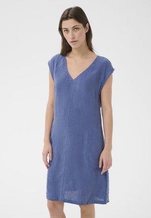 Femeie cu păr castaniu drept, purtând o rochie albastră din in fără mâneci cu decolteu în V, stând în fața unui fundal simplu, deschis la culoare.