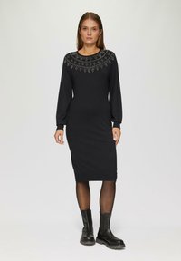 Schwarzes langärmliges Kleid mit einem gemusterten, pulloverartigen Halsausschnitt, figurbetontem Schnitt und einfarbiger Saum, kombiniert mit schwarzen Stiefeletten.