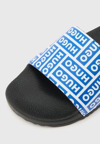 Μαύρο σαγιονάρες με υφή και φαρδιά λευκή λουριά διακοσμημένη με μπλε σχέδιο που επαναλαμβάνει τα λογότυπα "HUGO" καλύπτοντας το πάνω μέρος.