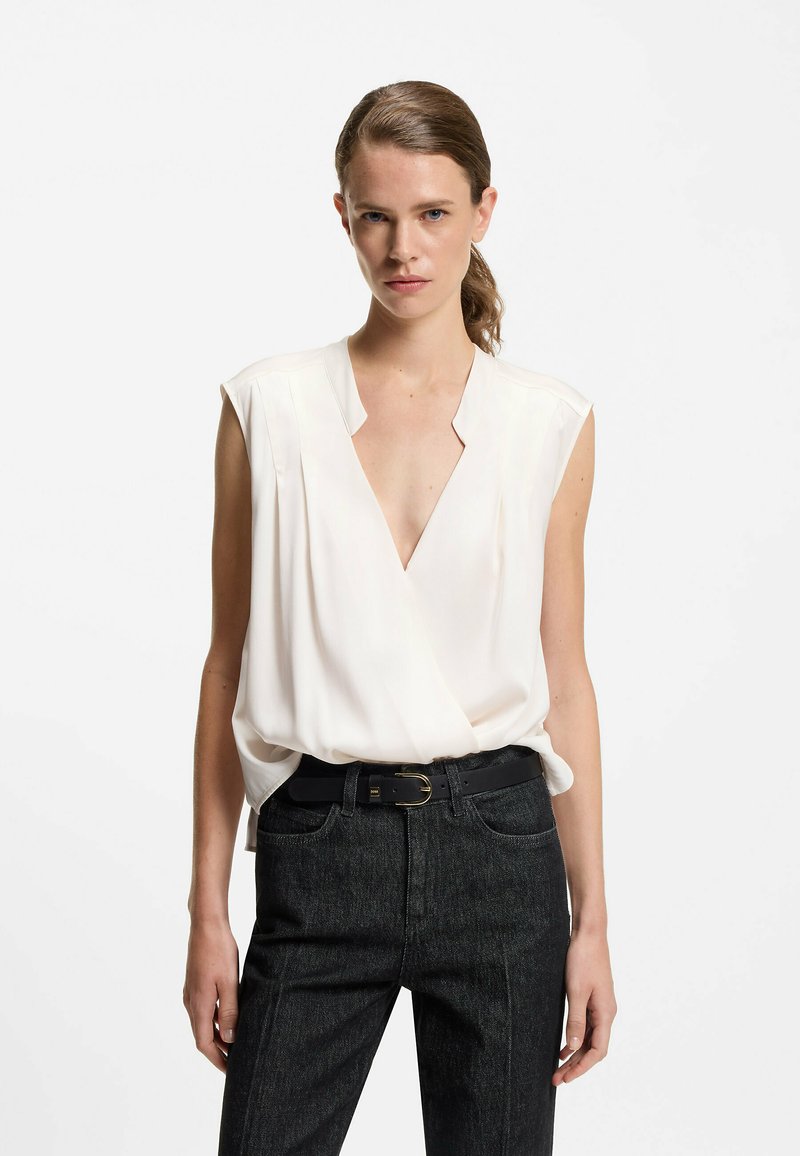 Weißes ärmelloses Oberteil mit tiefem V-Ausschnitt, weicher Textur und Faltenoptik. Kombiniert mit dunklen, hochgeschnittenen Jeans und einem schwarzen Gürtel.